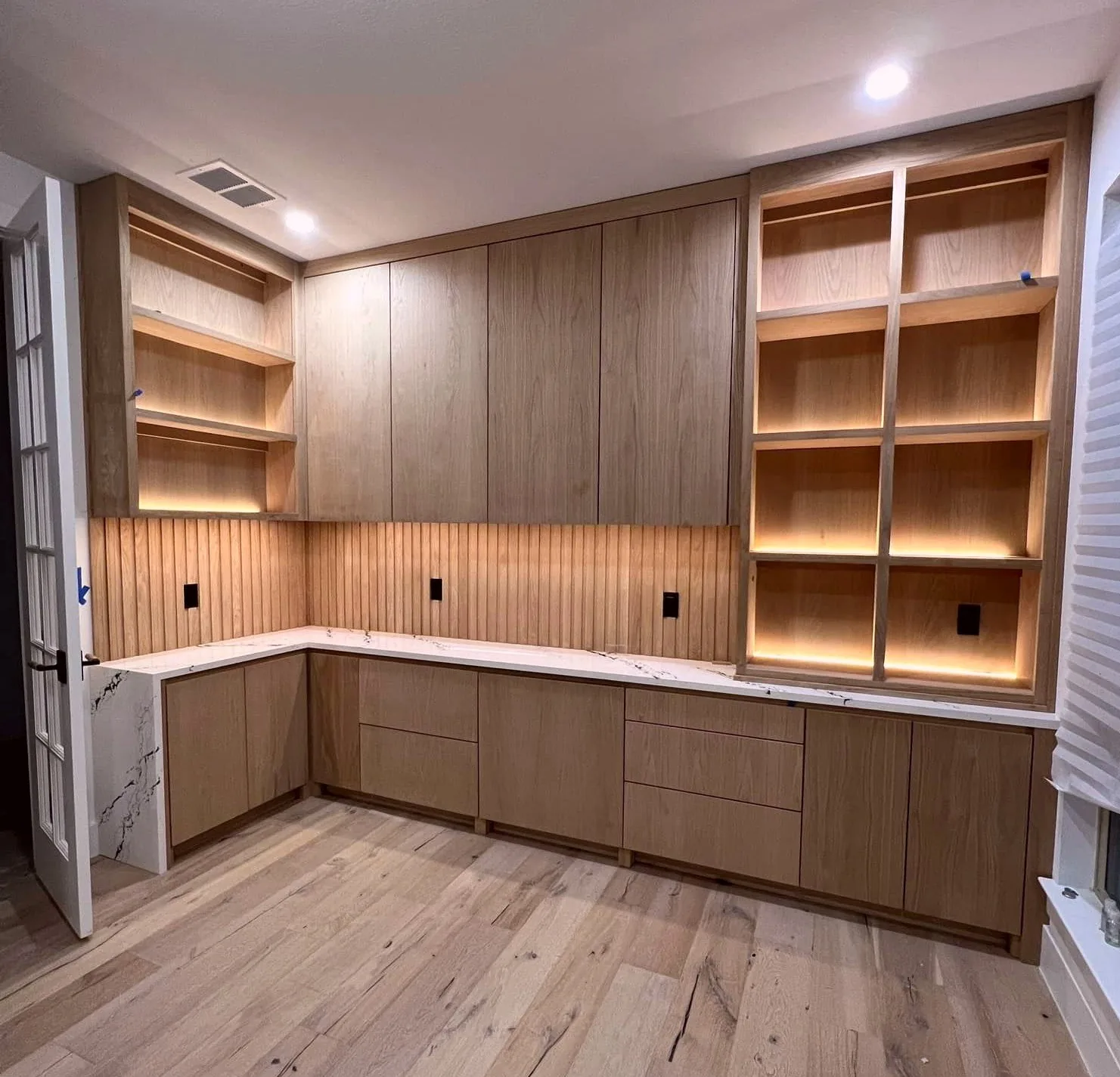 White oak built-in wall with lit open shelving, a run of base cabinets, and a stone counter